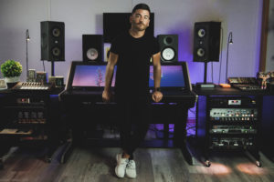 a man stands facing the camera as he leans against sound boards in his recording studio