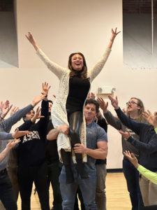 The lead actress spreads her arms victoriously as she is held aloft by a group of male chorus members