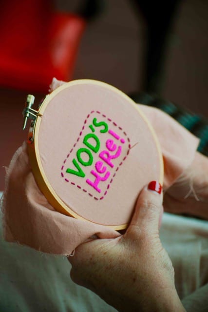 a needlepoint circle with the Vod's Here logo rendered in green and magenta