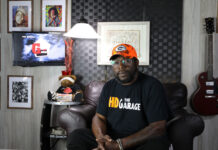 Listen Hear: HD The Garage- Toledo’s Video Podcast Covering Music, Culture & Untold Stories A man in a room with pictures and a guitar hanging in the background.