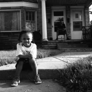 A little girl sitting on the sidewalk outside a house with a porch. On the porch an older person is sitting.